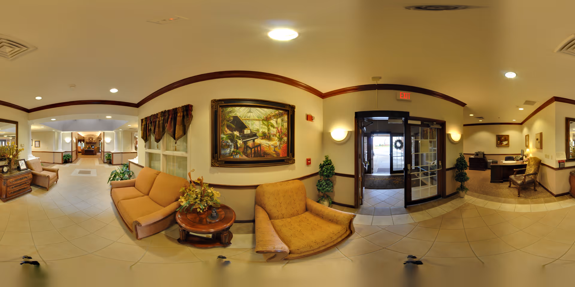 A spacious and warmly decorated senior living facility lobby with beige tiled floors and cream-colored walls accented by dark wood trim. The seating area includes cushioned armchairs and sofas in soft brown tones, arranged around a small wooden table with a floral arrangement. A large framed painting of a piano and garden scene hangs on the wall above one sofa. To the right, glass double doors lead outside, and to the far right is a reception desk with chairs and framed artwork on the walls. The hallway extends to the left with additional seating and plants along the way.