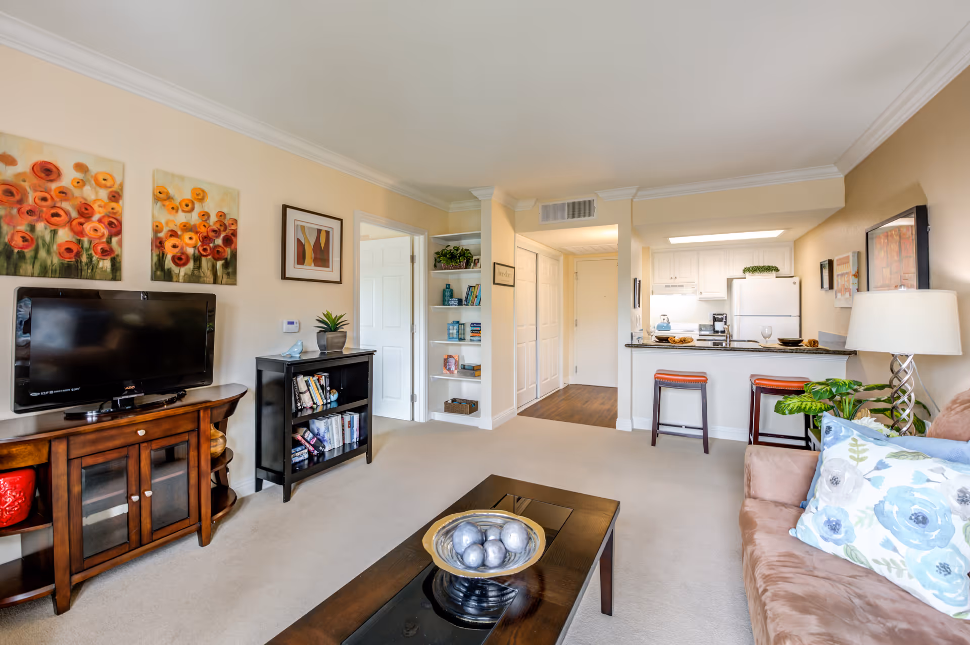 A bright and cozy living room area in a senior living facility with beige walls and carpet. The room features a brown sofa with floral cushions, a wooden coffee table with a decorative bowl, a TV on a wooden stand, and a small black bookshelf with books and decor. The room opens into a kitchen area with white cabinets, a refrigerator, and a breakfast bar with two stools. Colorful floral paintings and framed artwork decorate the walls.