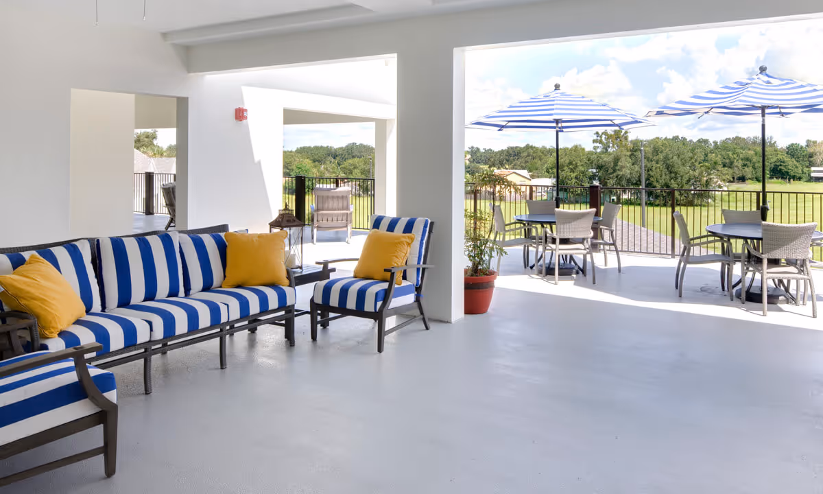Covered outdoor patio area with blue and white striped cushioned seating and yellow pillows, round tables with chairs and blue and white striped umbrellas, overlooking a green lawn and trees under a partly cloudy sky.