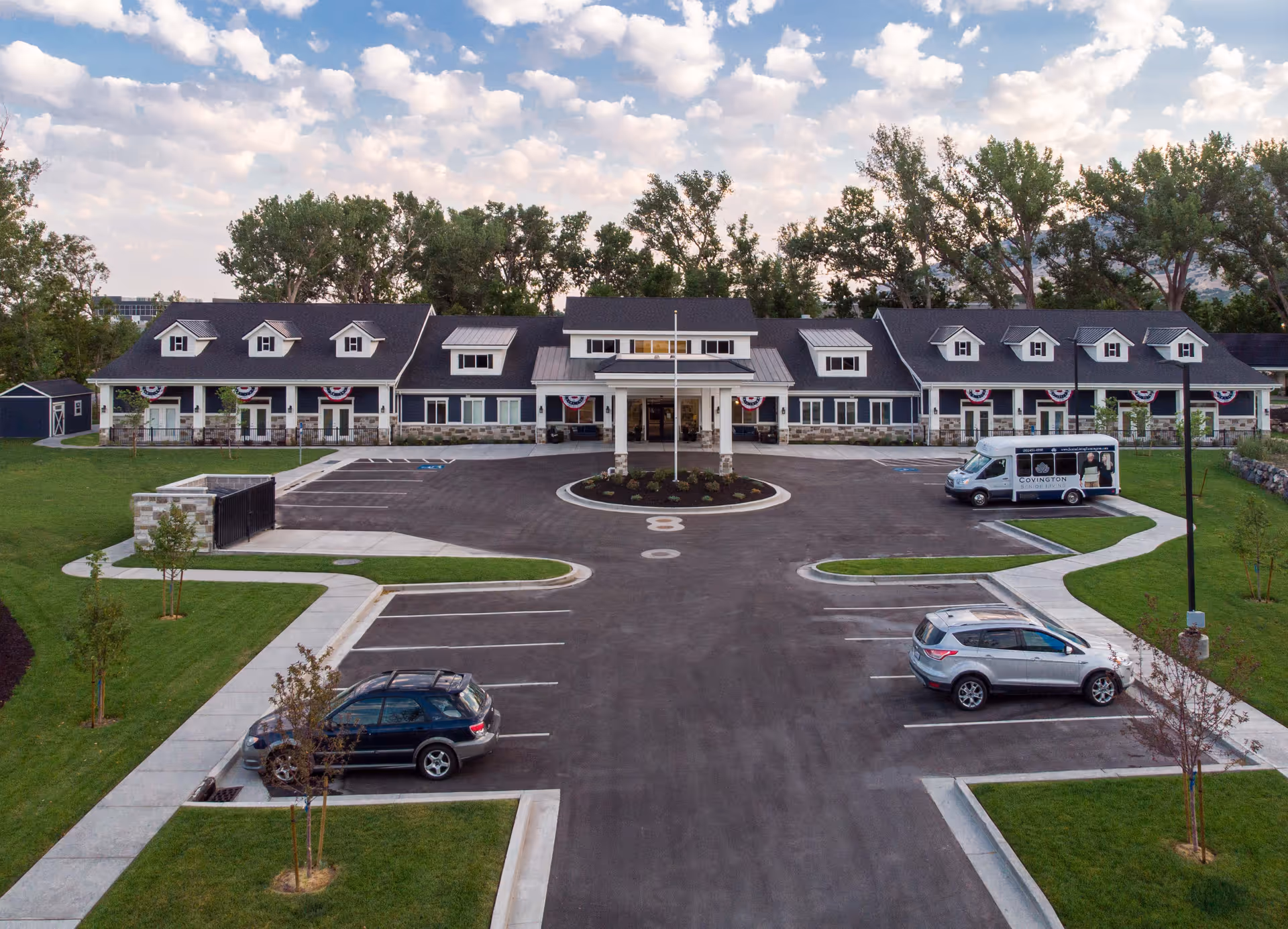 Wide frontal view of a single-story senior living building with a circular driveway, parked cars, and a shuttle van under a partly cloudy sky.