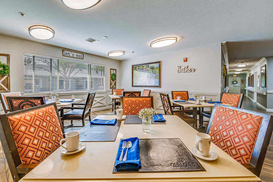 A bright dining room with several tables set with blue napkins, silverware, white mugs, and small flower arrangements. The chairs have orange patterned upholstery. The walls are decorated with framed pictures and signs that say 'let's eat' and 'gather'. Large windows with blinds allow natural light into the room.