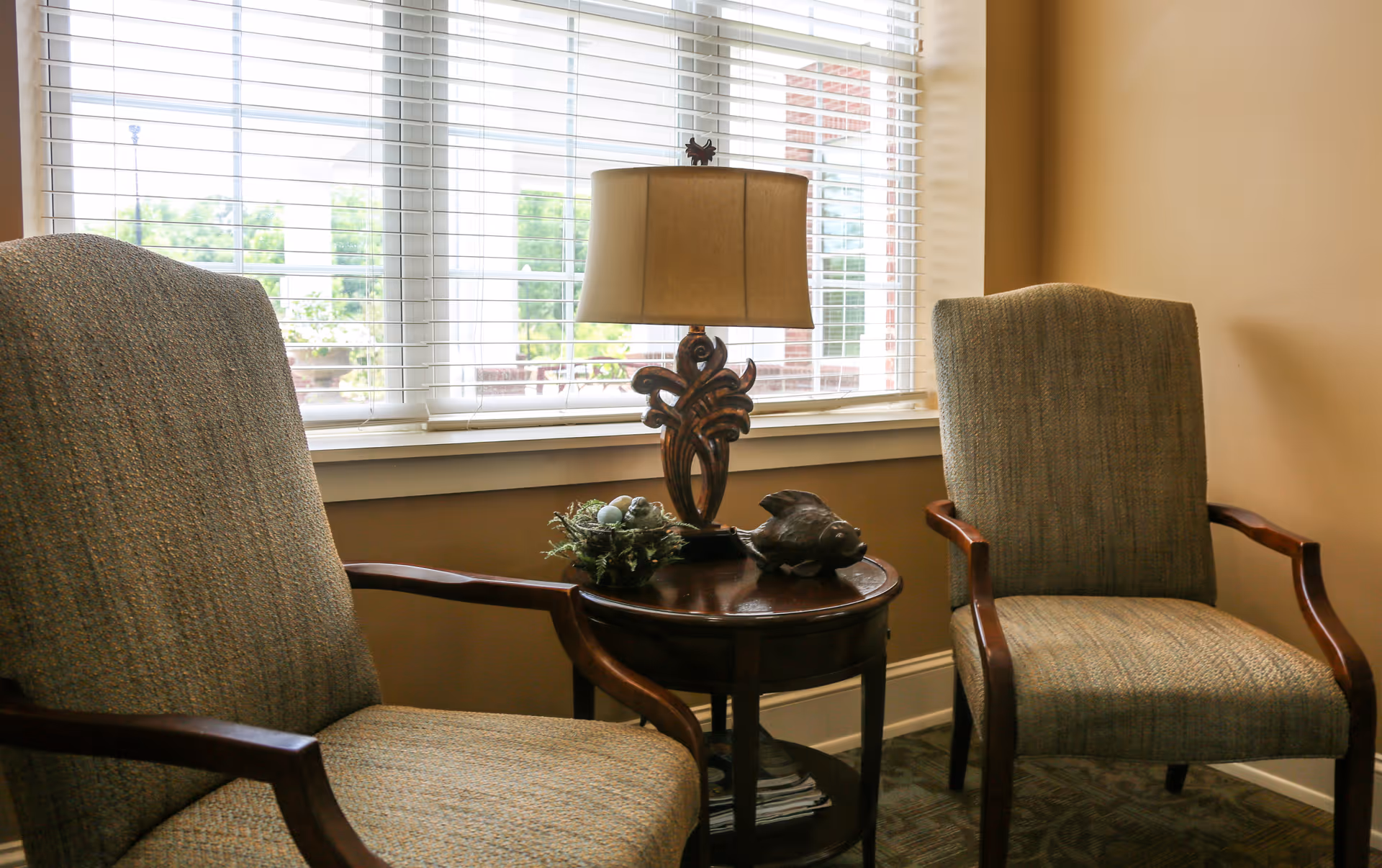 Two upholstered armchairs with wooden arms positioned on either side of a round wooden side table. On the table is a decorative lamp with a beige shade, a small plant arrangement, and a fish-shaped ornament. Behind the chairs is a window with white blinds partially open, allowing natural light to fill the room.