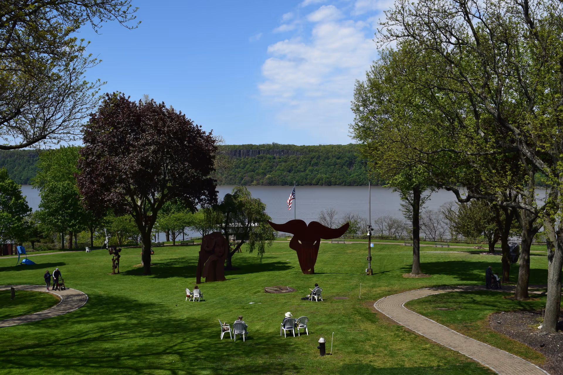 A scenic outdoor park area with green grass, several trees, and a paved walking path. There are metal sculptures of an elephant and a bull's head in the center. People are seated on white chairs scattered around the lawn, and a large body of water with a tree-covered hill in the background is visible under a partly cloudy sky.