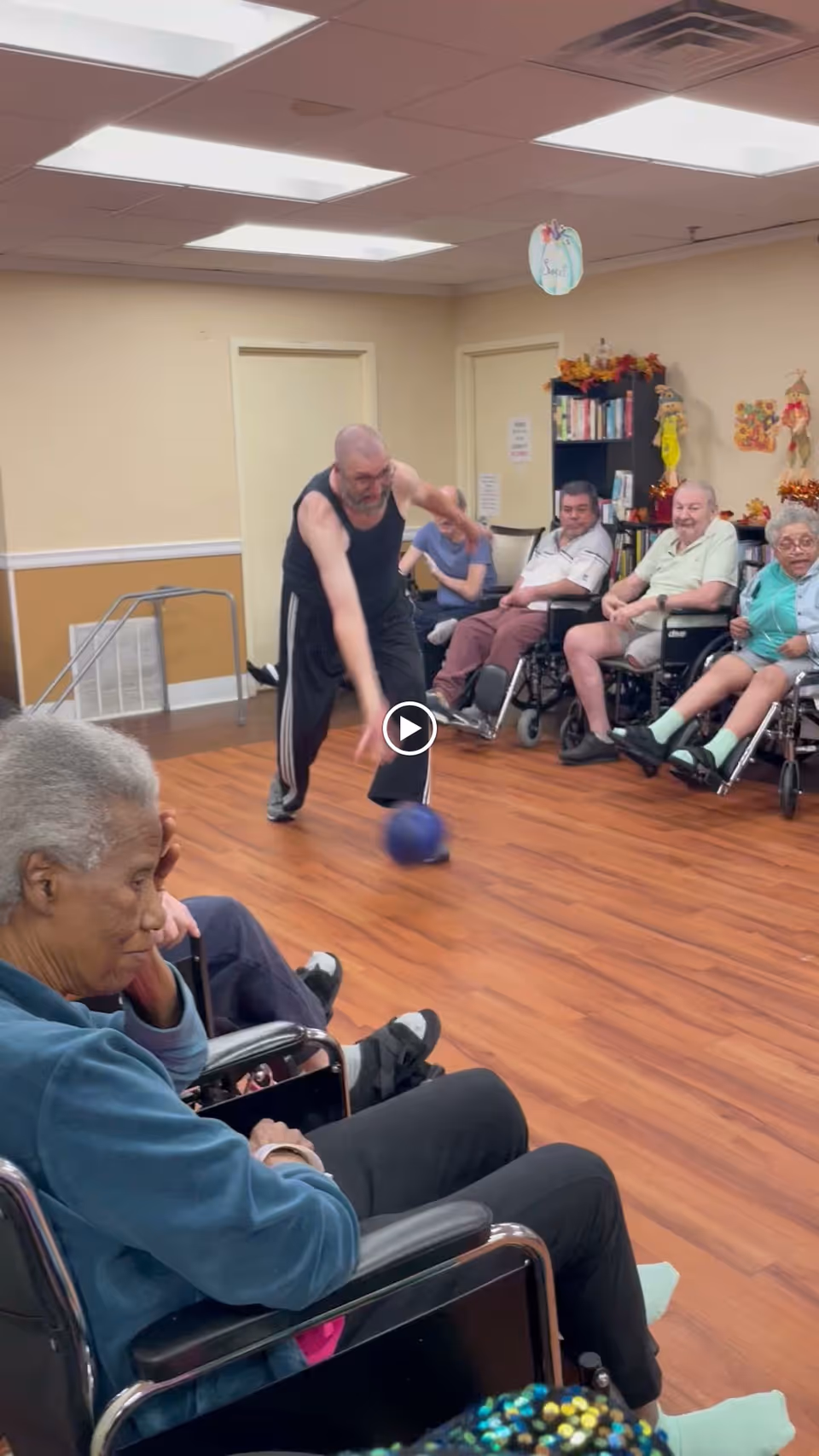 A group of elderly people sitting in wheelchairs arranged in a semicircle in a room with wooden flooring. One man in a black tank top and pants is actively rolling a blue ball on the floor, engaging in a group activity. The room has beige walls, fluorescent ceiling lights, and a bookshelf with decorations in the background.