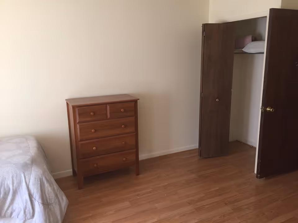 A simple bedroom with a wooden floor, a wooden chest of drawers, a partially visible bed with a light-colored bedspread, and an open closet with wooden doors showing a pillow and blanket inside.