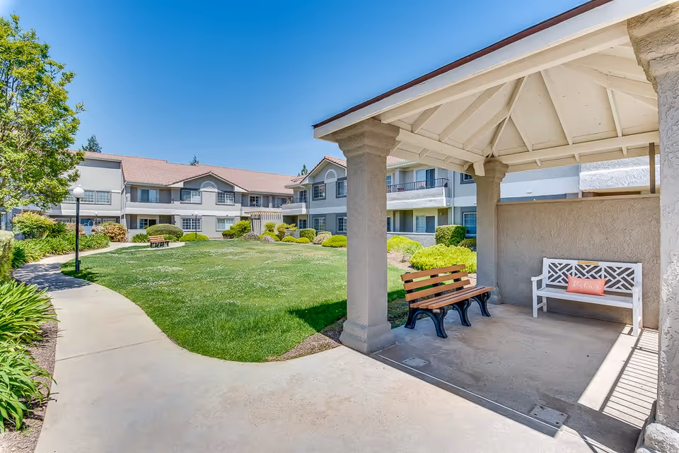 Outdoor courtyard area of a senior living facility with a covered seating area featuring two benches, one with a pink pillow that says 'relax'. The courtyard has a well-maintained lawn, shrubs, and a paved walkway. The building surrounding the courtyard is two stories with balconies and windows under a clear blue sky.