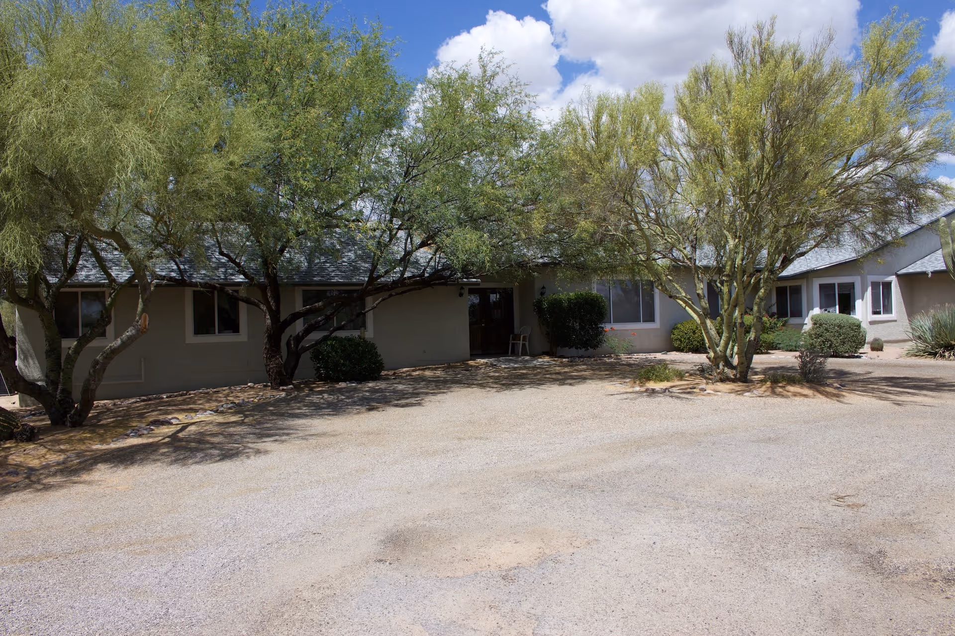 Exterior view of a single-story building with a gravel driveway and several trees in front under a partly cloudy sky.