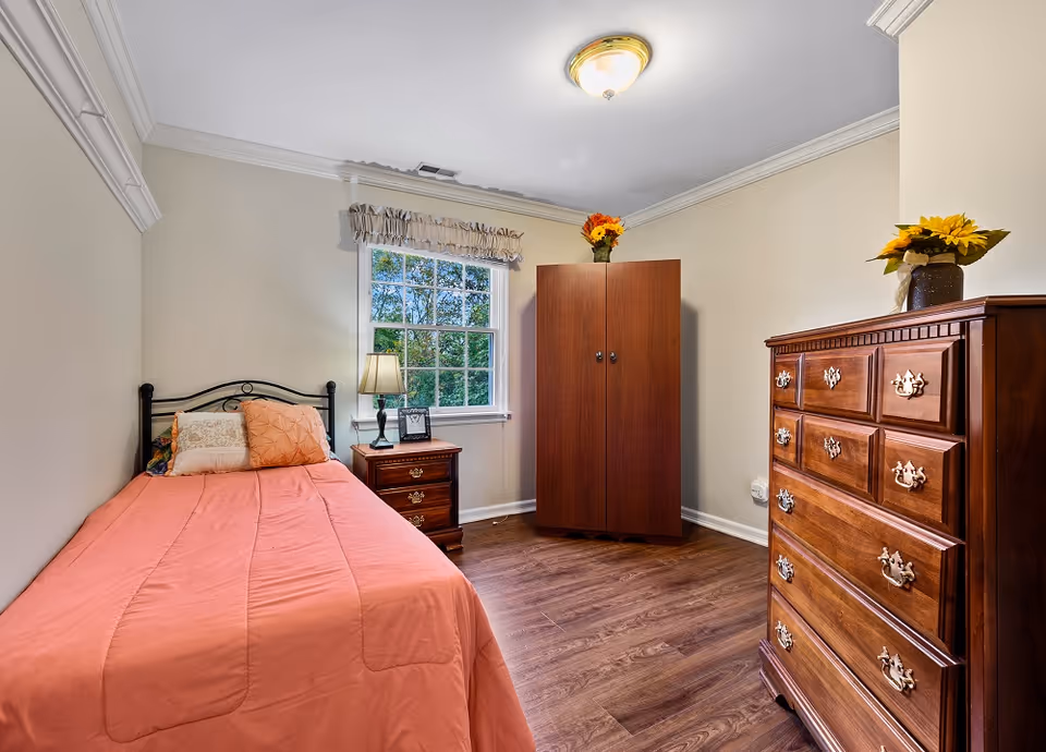 A small, cozy bedroom with a single bed covered in a coral-colored comforter and multiple pillows. Next to the bed is a wooden nightstand with a lamp and a framed photo. Across from the bed is a wooden dresser with a vase of sunflowers on top, and a wooden wardrobe with a vase of flowers on it is positioned near a window with a beige valance. The room has light-colored walls and wood flooring.