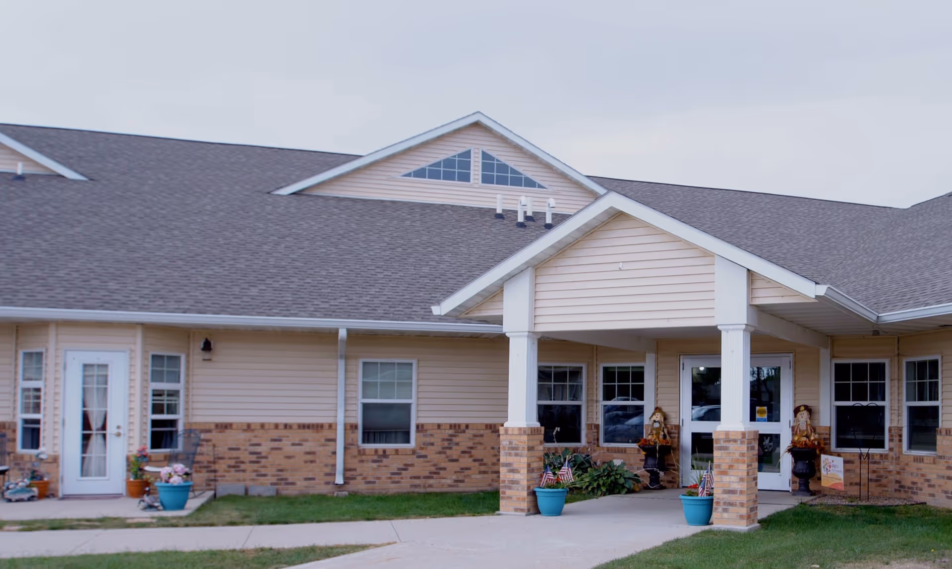 Exterior view of a single-story building with beige siding and brick accents, featuring a covered entrance supported by white columns. There are several windows and a glass door, with potted plants and seasonal decorations near the entrance.