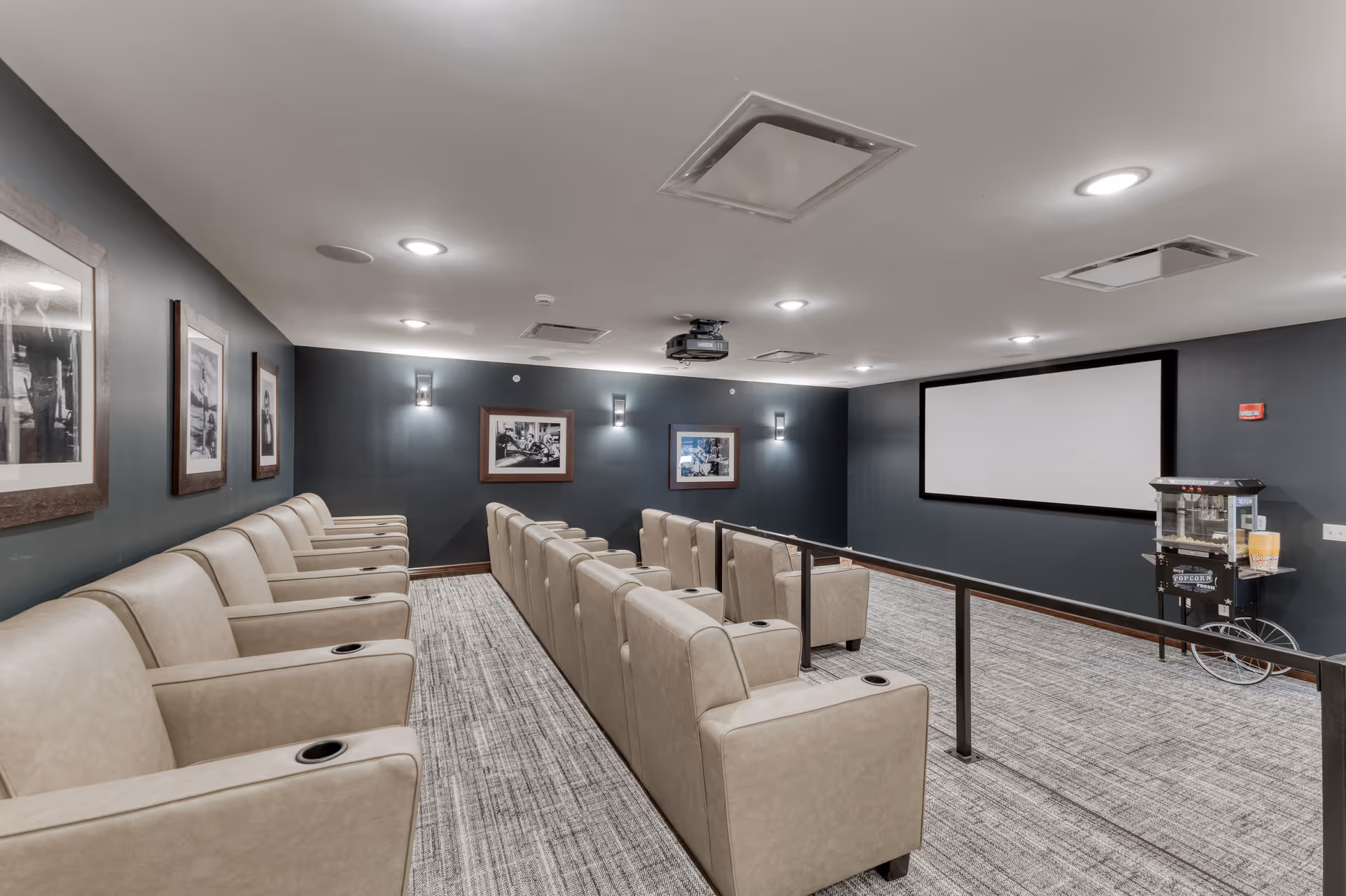 A small movie theater room with two rows of beige reclining chairs facing a large blank projection screen. The walls are painted dark gray and decorated with framed black and white photographs. A popcorn machine is positioned near the screen on the right side. The ceiling has recessed lighting and a projector mounted in the center.