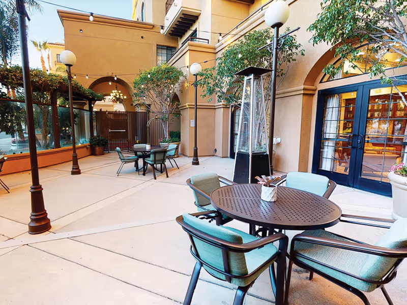 A sunlit outdoor courtyard with round metal tables, cushioned chairs, tall lamps, and potted trees in front of building entrances.