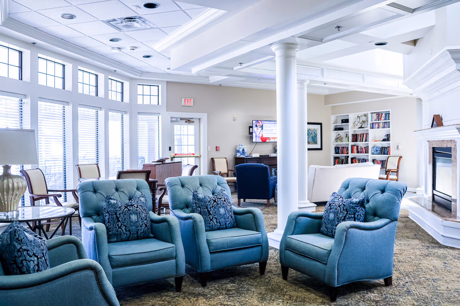 A bright senior living lounge with several blue upholstered armchairs arranged around a column, fireplace, and bookshelves near large windows.