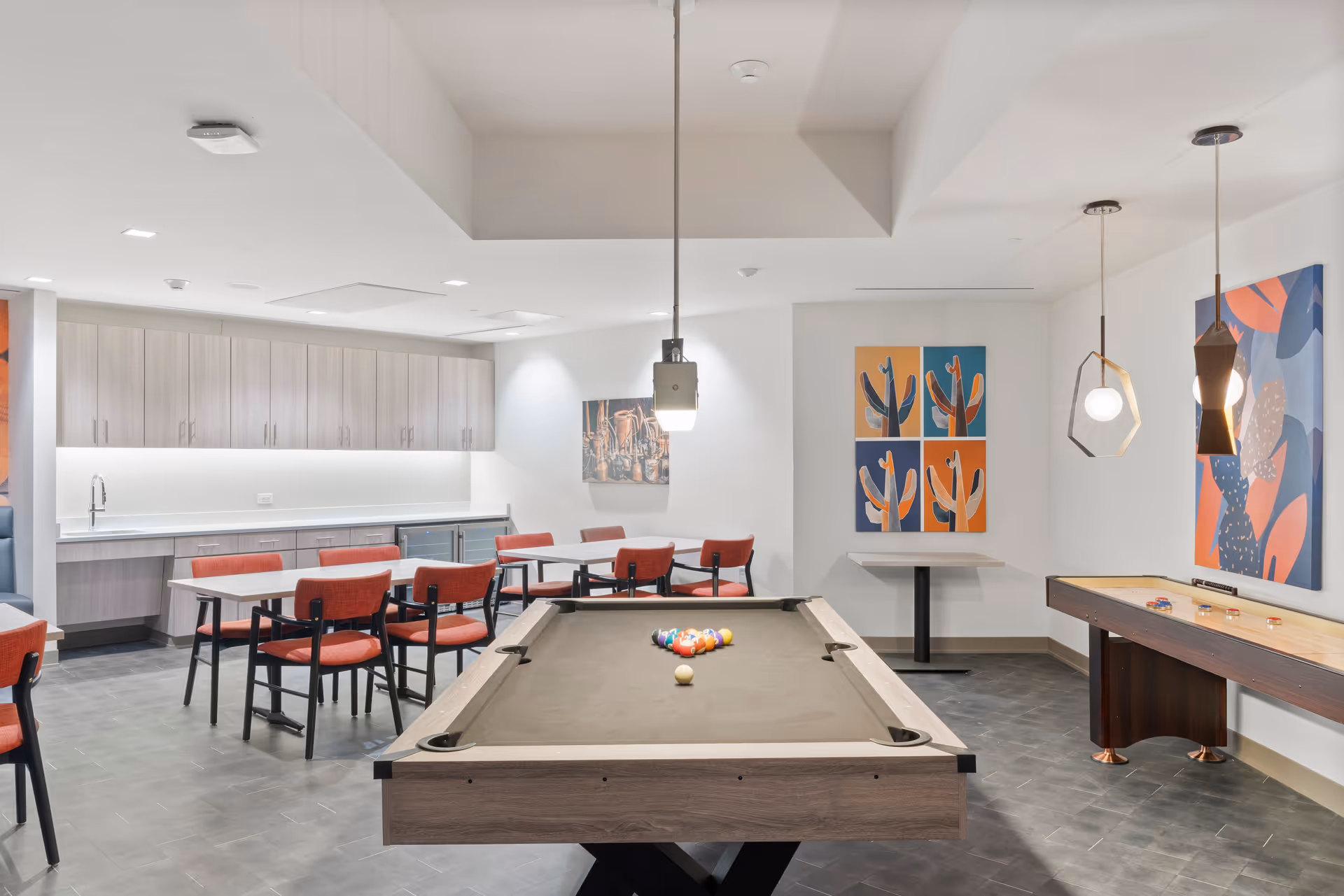 A recreational room featuring a pool table with balls arranged for a game, a shuffleboard table, several red cushioned chairs around tables, modern pendant lights hanging from the ceiling, colorful abstract artwork on the walls, and a kitchenette area with cabinets and a sink in the background.