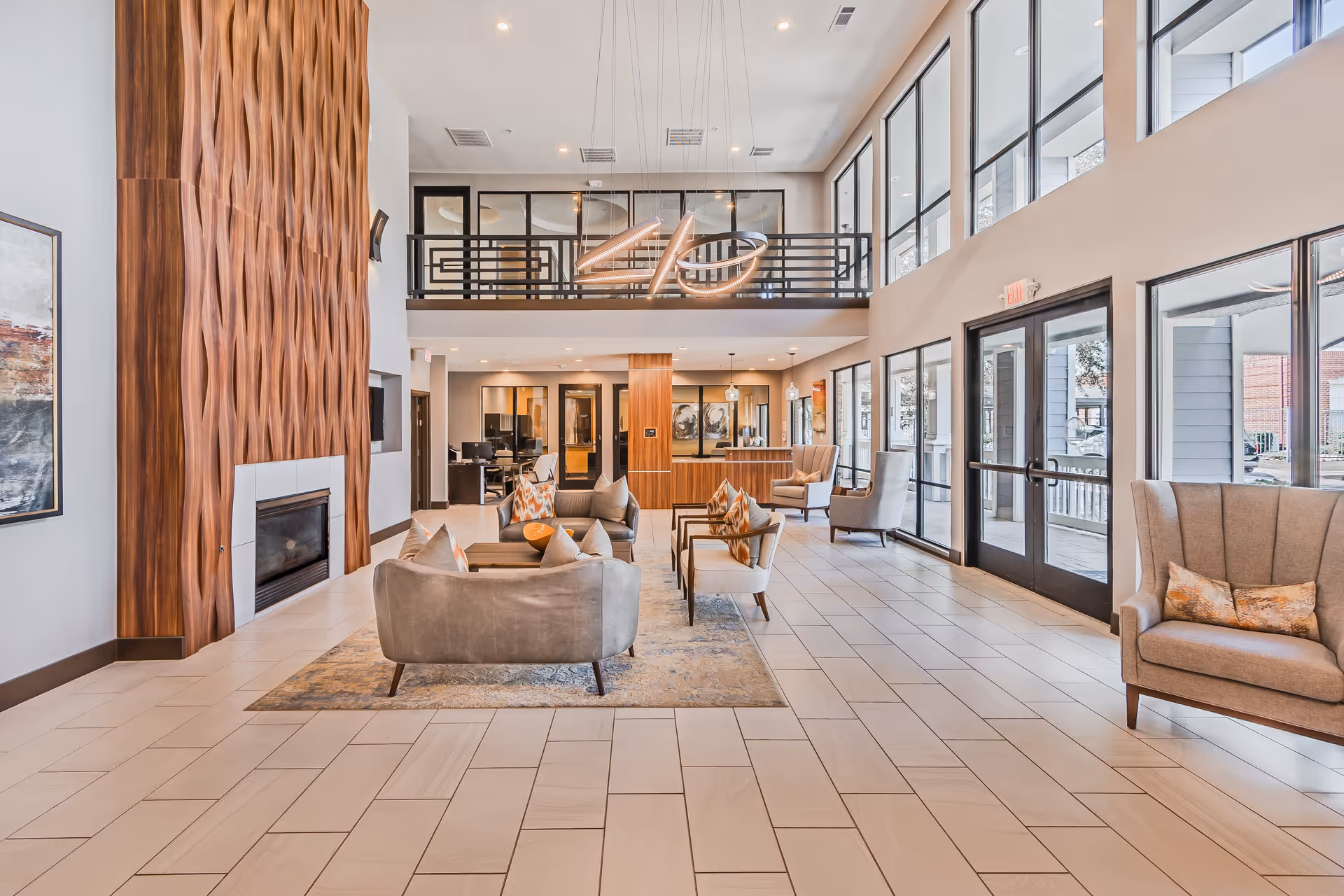 Spacious and modern senior living facility lobby with high ceilings, large windows, and a seating area featuring sofas and armchairs around a coffee table. A decorative wooden fireplace is on the left wall, and a reception desk is visible in the background. The space is well-lit with natural light and contemporary light fixtures.