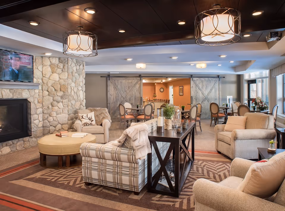 Bright, cozy common living area with a stone fireplace, armchairs and sofas around a central table and dining tables beyond.