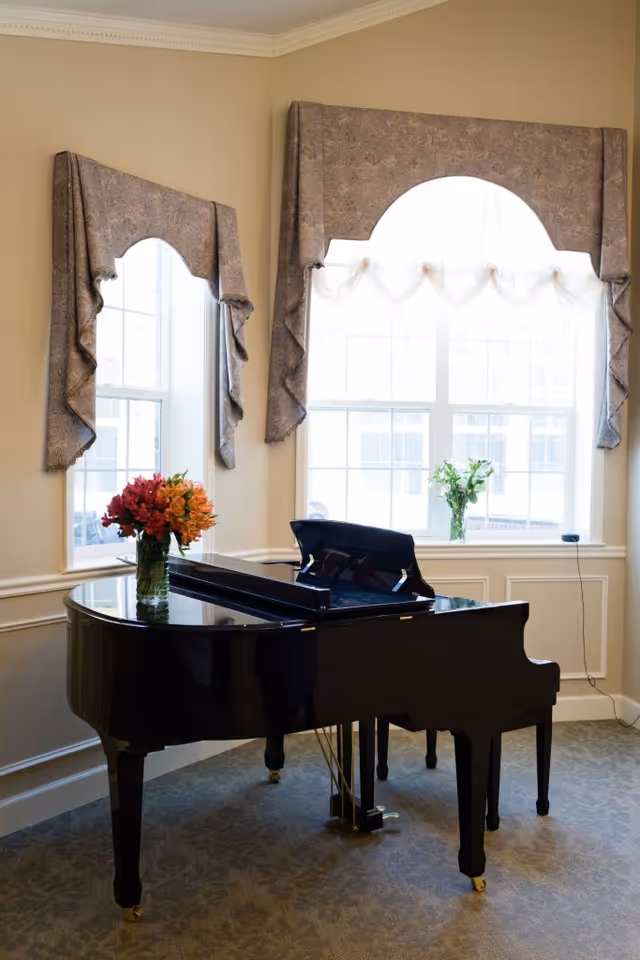 A black grand piano with a vase of colorful flowers on top is placed in a carpeted room with beige walls. Two large windows with decorative valances allow natural light to fill the space, and a small vase with green foliage is on the windowsill.