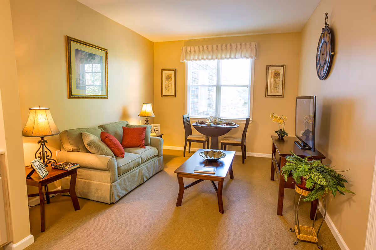 A cozy living room with a green sofa adorned with red and green pillows, two wooden side tables each with a lamp, a wooden coffee table with a decorative bowl, a small round dining table with two chairs near a window, a TV on a wooden stand, a large wall clock, framed artwork on the walls, and a potted plant on a stand.