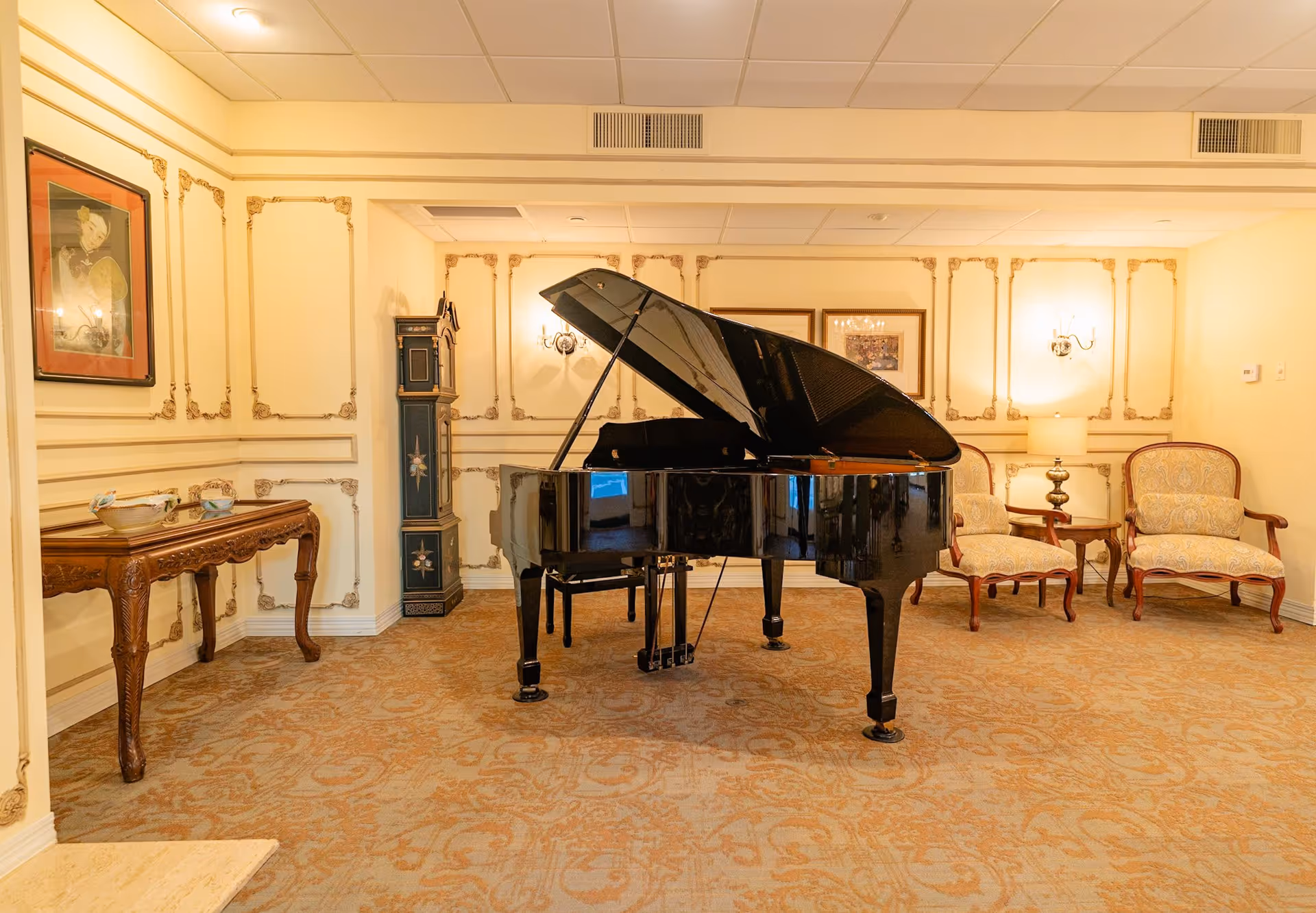 A cozy interior room featuring a black grand piano in the center, two upholstered armchairs with wooden frames, a wooden side table with decorative bowls, a tall grandfather clock, framed artwork on the walls, and warm lighting from wall sconces and a table lamp.