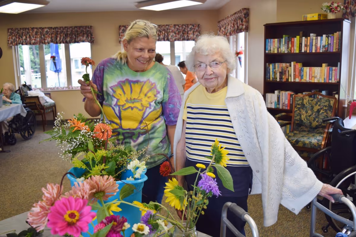 Two women, one elderly and one middle-aged, standing together indoors near a table with colorful flowers in vases. The elderly woman is using a walker and wearing a white cardigan over a striped shirt. The middle-aged woman is smiling and holding a flower, wearing a tie-dye shirt. In the background, there are other people seated, a bookshelf filled with books, and windows with floral curtains.