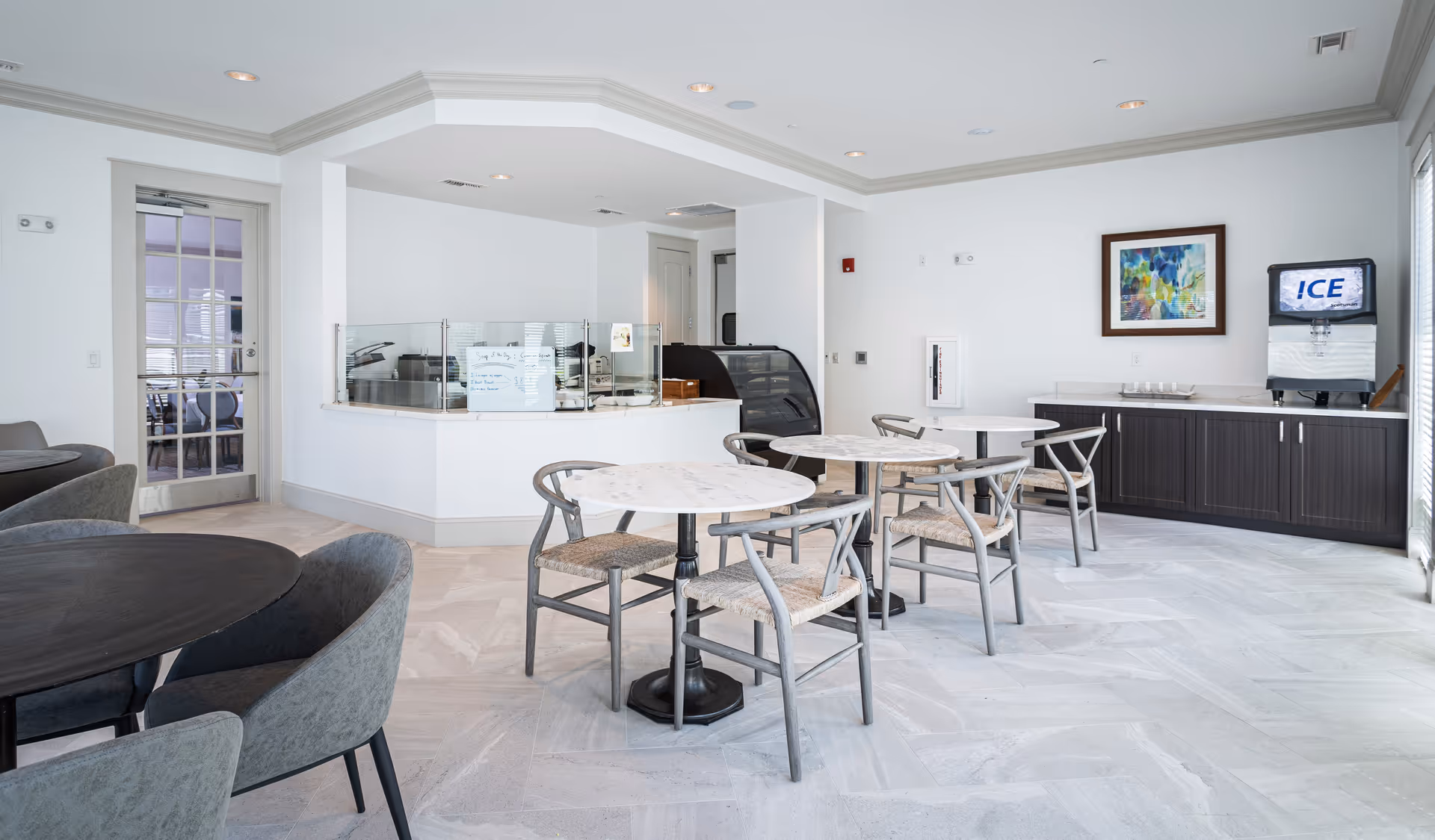 A bright dining area with round marble-top tables and wooden chairs with woven seats. There is a counter with a glass sneeze guard, a black display case, and a whiteboard with menu items. On the right side, there is a cabinet with an ice dispenser and a colorful framed artwork on the wall. The room has light-colored tile flooring and white walls with crown molding.