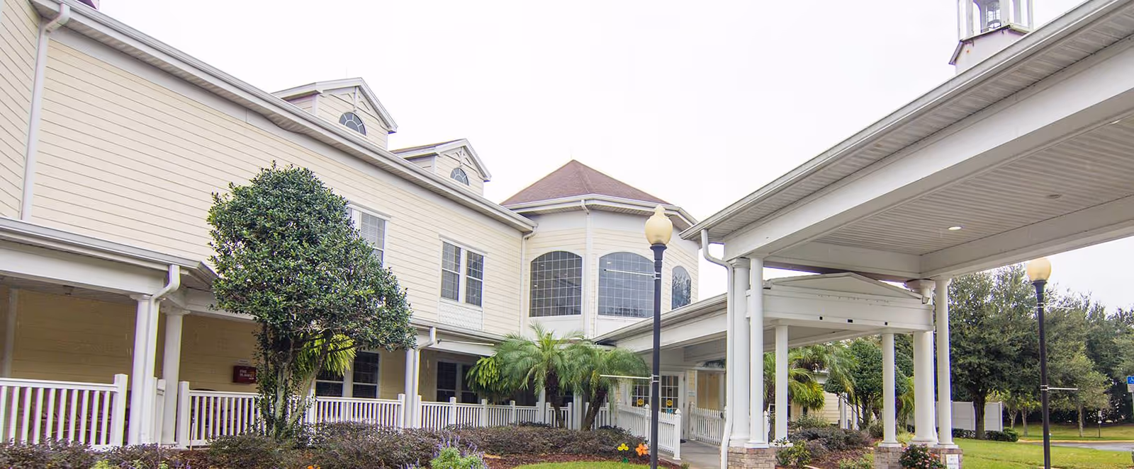 Exterior view of a senior living facility named Madison at Ocoee, showing a two-story building with beige siding, large windows, a covered entrance with white columns, landscaped greenery including trees and bushes, and lamp posts along the walkway.