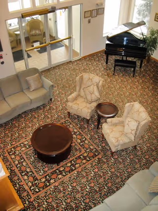 Overhead view of a senior living facility common area with floral patterned carpet, two beige upholstered armchairs with matching pillows, a round wooden coffee table, a smaller round side table, two sofas with cushions, and a black grand piano near large windows and glass doors.