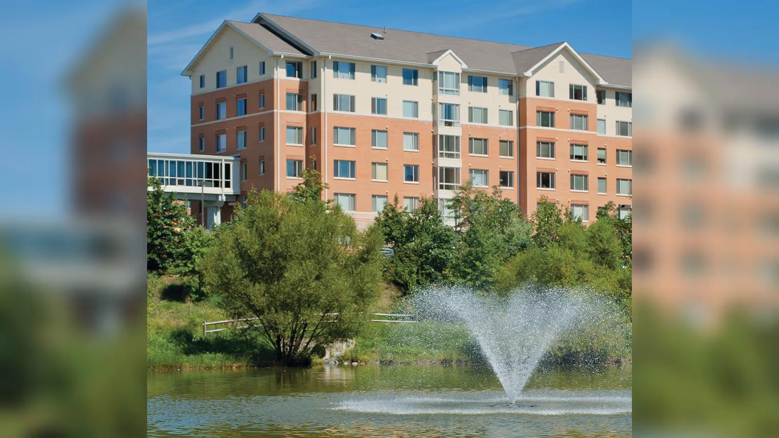 A multi-story senior living building with a pond, trees, and a fountain in front.