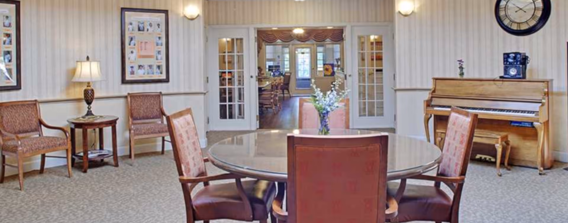 Communal sitting area with a round table and chairs, additional seating, a piano, and glass double doors leading to another room.