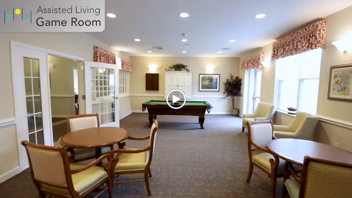 A spacious assisted living game room with a pool table in the center, round tables with chairs, and comfortable armchairs near windows with floral valances. The room has beige walls, carpeted floor, and bright ceiling lights.