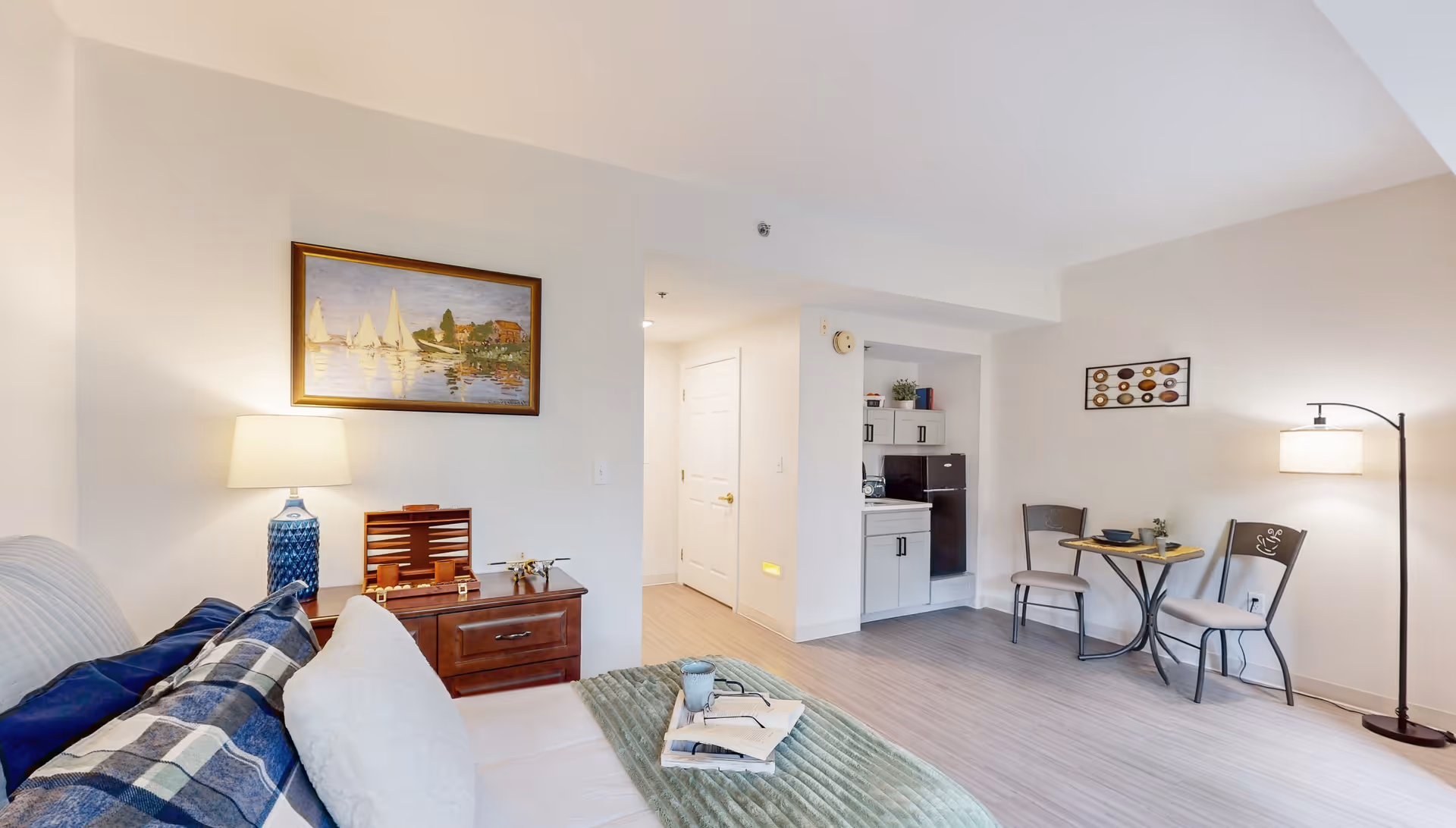 A cozy living space featuring a bed with pillows and a green throw blanket, a wooden nightstand with a blue lamp and decorative items, a small dining area with two chairs and a round table, and a kitchenette with white cabinets and a black refrigerator. The walls are light-colored with framed artwork, and the floor has light wood-style flooring.