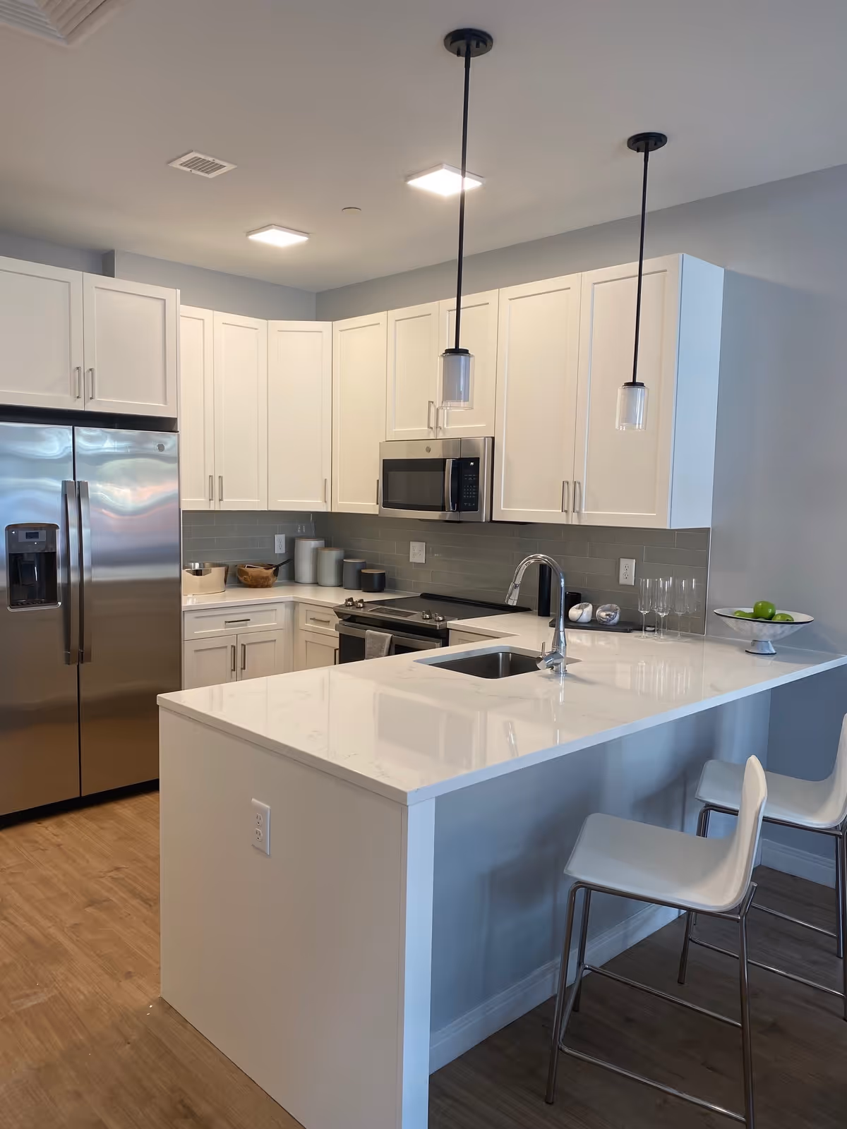 Modern kitchen with white cabinets, stainless steel appliances, a marble-topped island with sink and bar stools, and pendant lights.