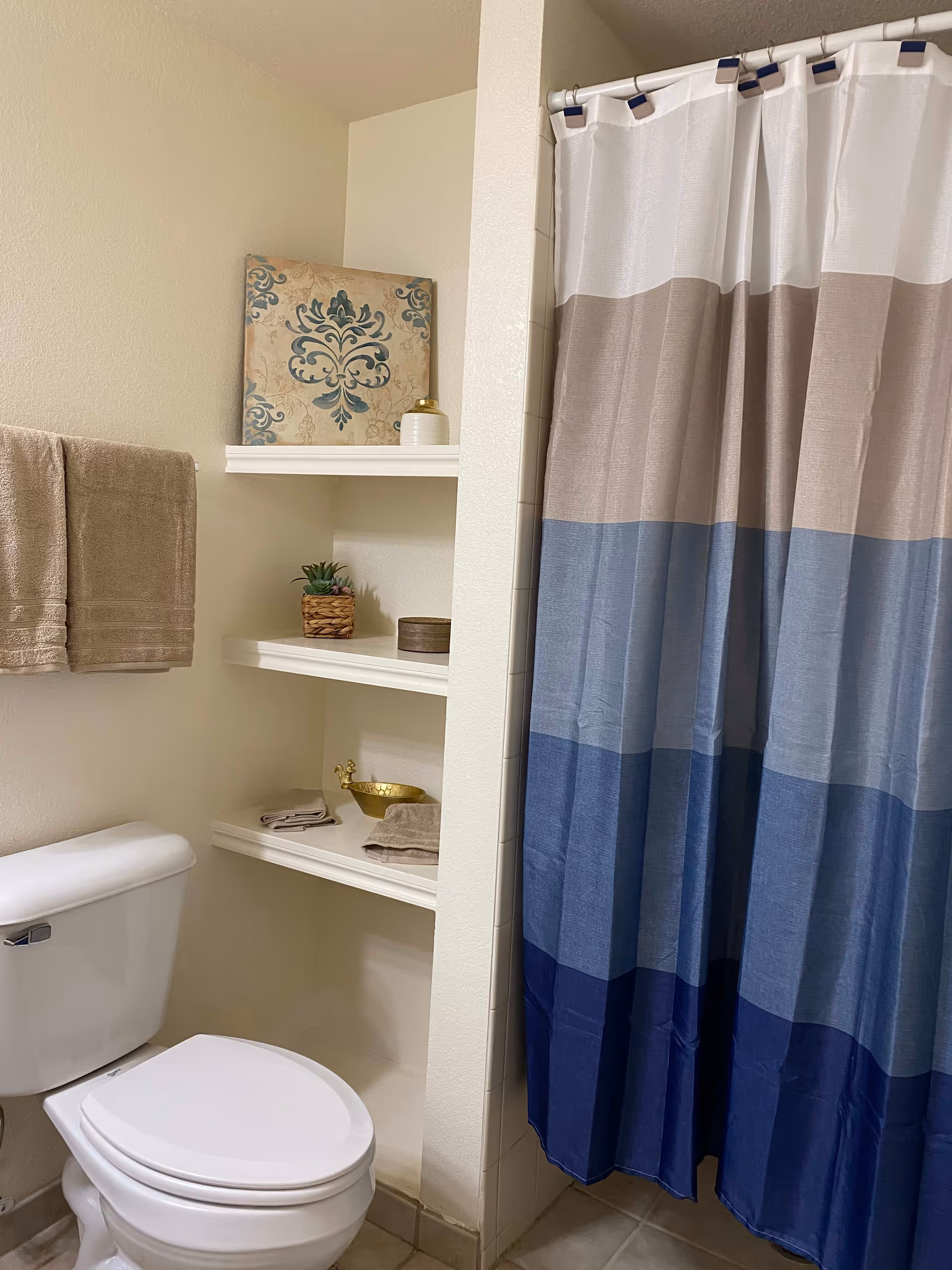 A bathroom with a white toilet, beige towels hanging on a wall-mounted towel rack, built-in white shelves holding decorative items including a small plant, a gold bowl, and a framed artwork. A shower with a curtain featuring horizontal stripes in white, beige, light blue, and dark blue is visible.