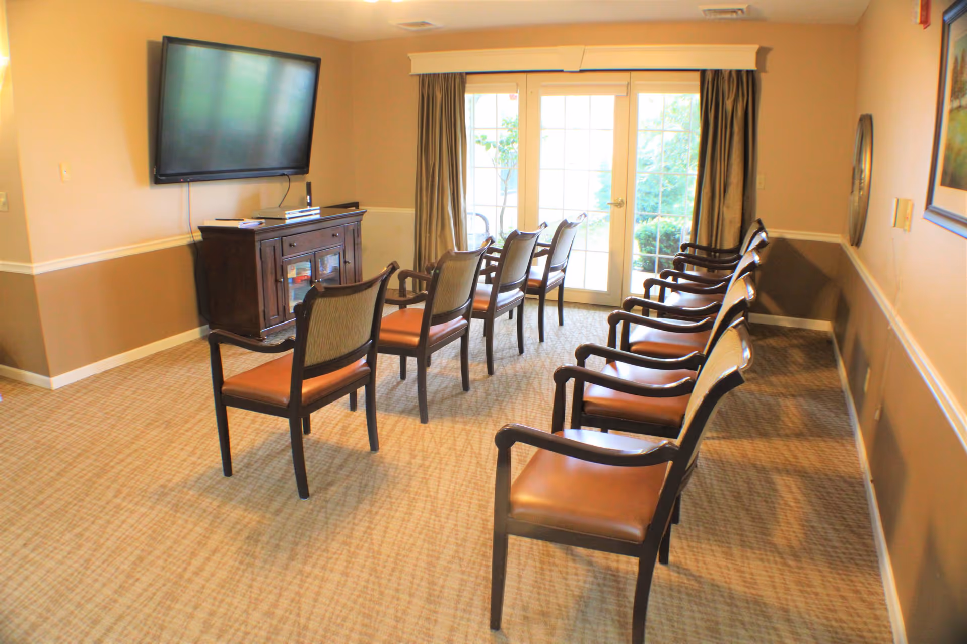 A small meeting or activity room with two rows of wooden chairs with brown cushions facing a wall-mounted flat screen TV above a wooden cabinet. The room has beige walls with white trim and a carpeted floor. At the far end, there are glass double doors with curtains, leading outside to a garden area.
