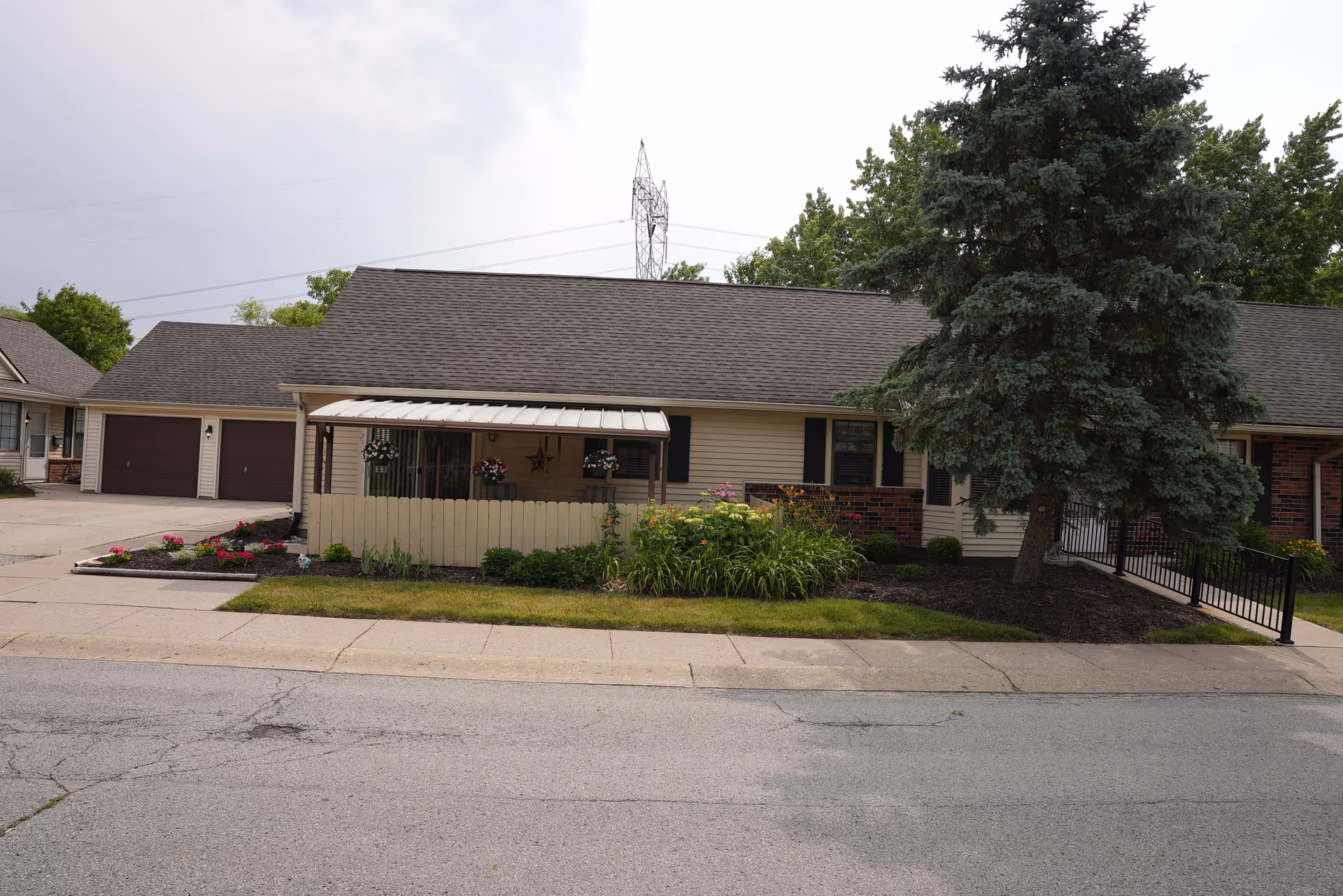 Single-story residential building with a covered porch, flower beds, and a large evergreen tree in front. The building has a beige exterior with dark brown shutters and a two-car garage on the left side. The scene is set on a paved street with a sidewalk.