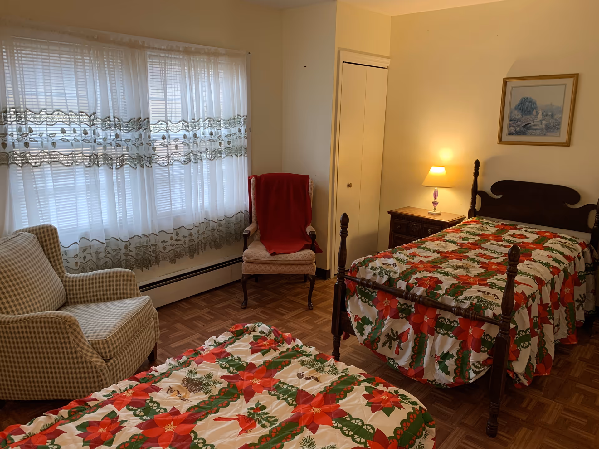 A cozy bedroom with two single beds covered in festive red and green floral bedspreads. There is a wooden nightstand with a small lamp between the beds. A framed picture hangs on the wall above one bed. Two armchairs, one with a red blanket draped over it, are placed near a window with sheer white curtains. The room has parquet flooring and light-colored walls.