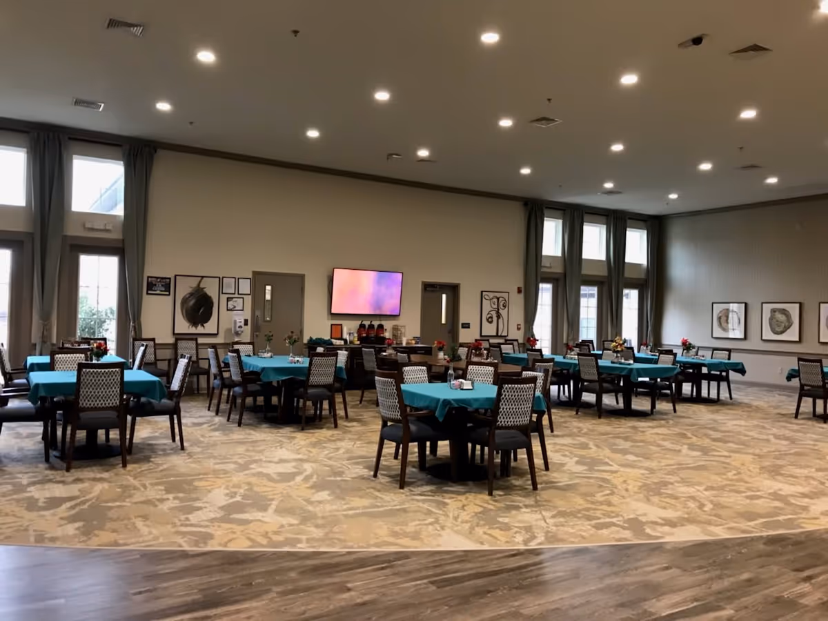 A spacious dining room with multiple tables covered in teal tablecloths and surrounded by chairs. The room has large windows with curtains, framed artwork on the walls, a patterned carpet, and a wooden floor section in the foreground. A flat-screen TV is mounted on the far wall above a counter with coffee dispensers and other items.