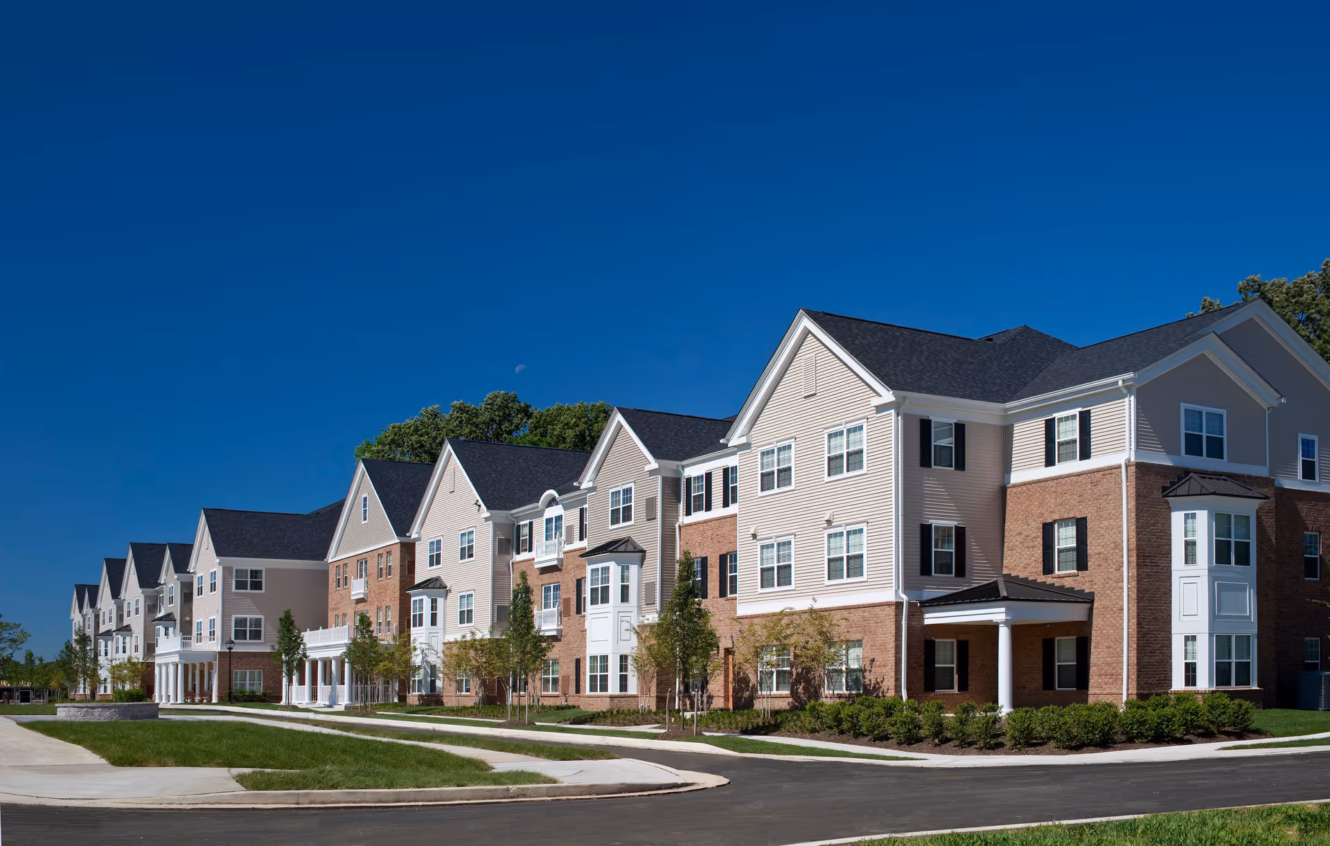 Exterior view of a large, modern senior apartment building with multiple stories, beige siding, brick accents, black shutters, and a dark roof under a clear blue sky.