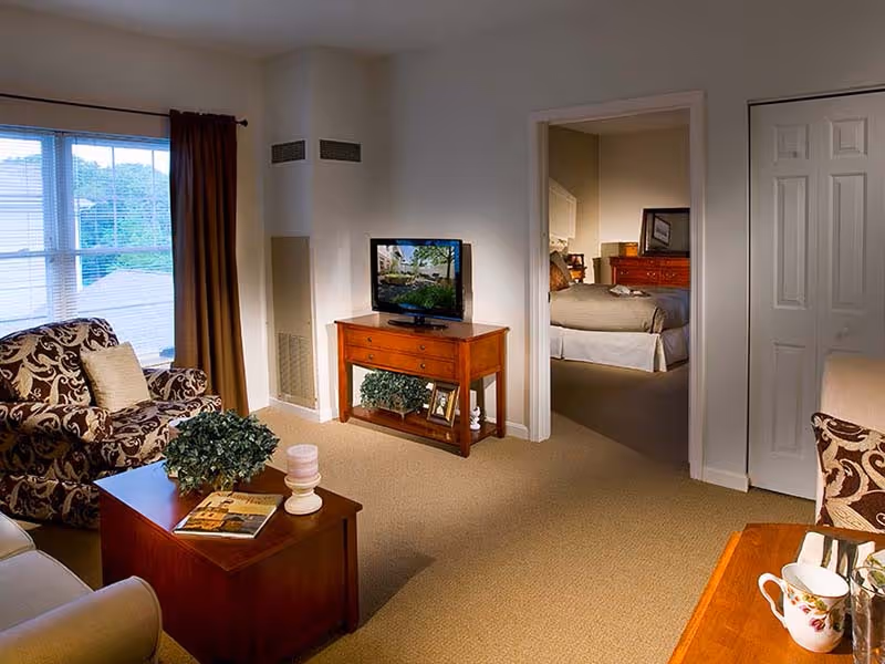 A cozy living room with patterned armchairs, a wooden coffee table and TV stand, and a doorway leading to a bedroom.