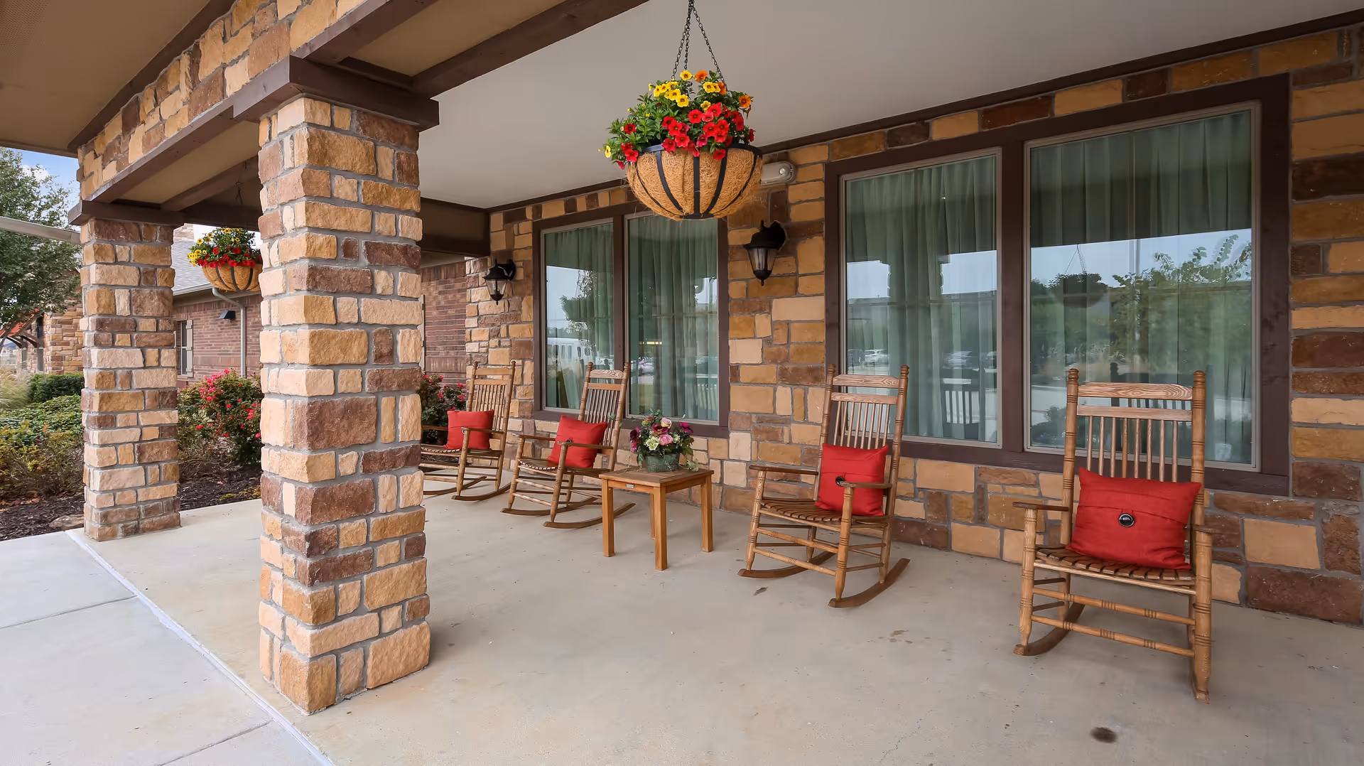 Covered outdoor patio area with stone pillars and walls, featuring wooden rocking chairs with red cushions and a small wooden table with a flower arrangement. Hanging flower baskets are suspended from the ceiling, and large windows with curtains are visible on the building wall.