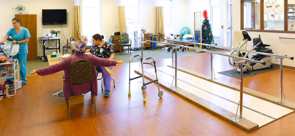 A rehabilitation or physical therapy room in a senior living facility with two women seated and exercising with yellow resistance bands. A walker is positioned nearby, and parallel bars are installed for walking support. A staff member in scrubs is organizing supplies on the left side. The room has wood flooring, exercise equipment, a Christmas tree, and large windows letting in natural light.