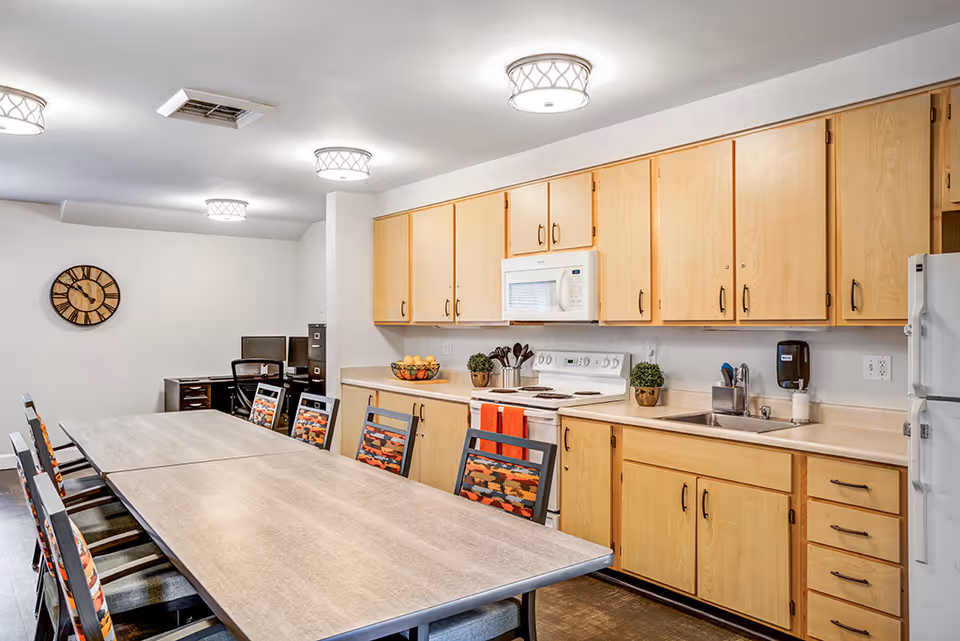 Communal dining area with a long table and chairs adjacent to a kitchen with light wood cabinets, stove, microwave, sink, and refrigerator.