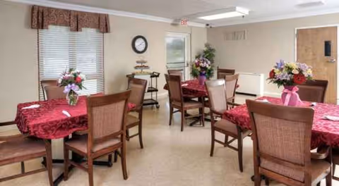 Dining room with round tables, red tablecloths, flower centerpieces, and chairs in a senior facility.