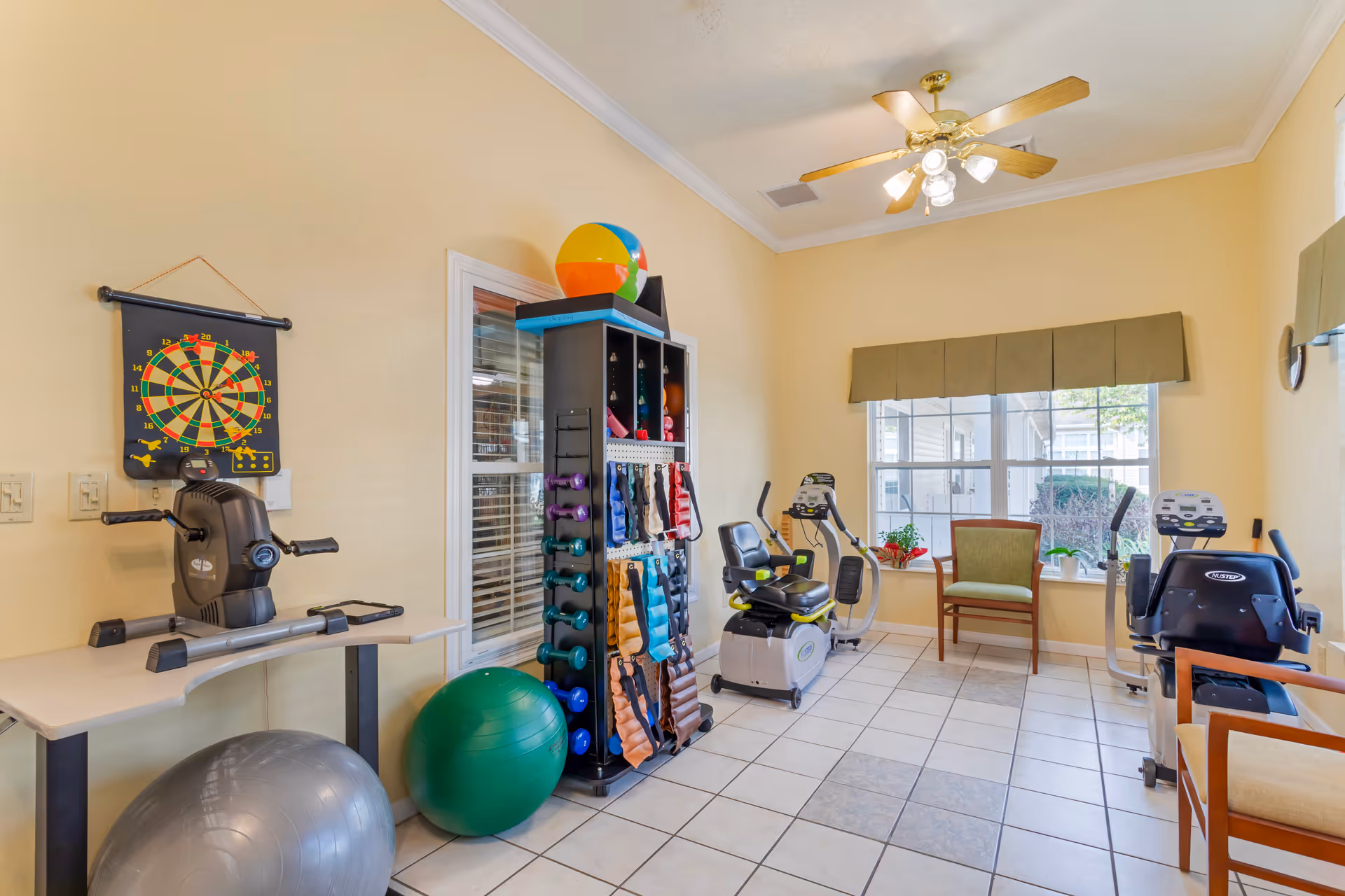 A small exercise room with tiled floor and beige walls, featuring two stationary recumbent bikes, a rack with various dumbbells and exercise bands, two large exercise balls, a dartboard on the wall, and a ceiling fan with lights. There are two chairs near a window with a green valance, letting in natural light.