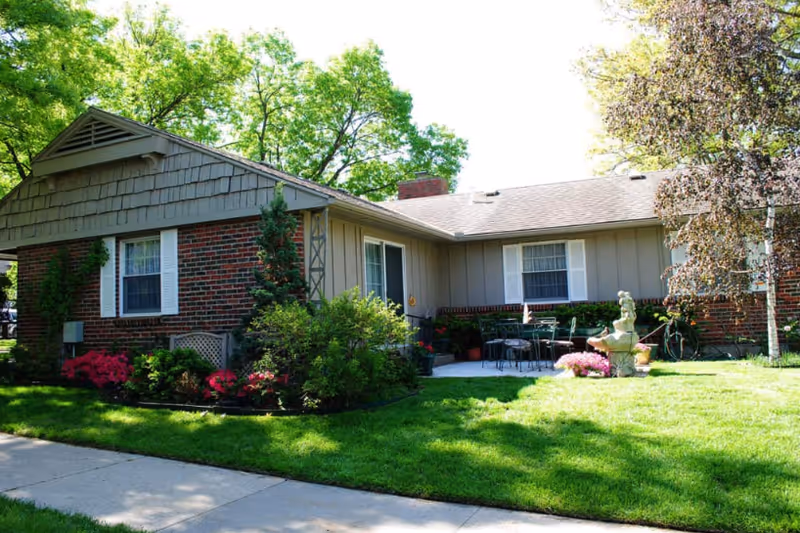 Single-story residential building with a brick and wood exterior surrounded by green grass, bushes, and trees. There is a small patio area with outdoor furniture and a decorative fountain in the front yard.