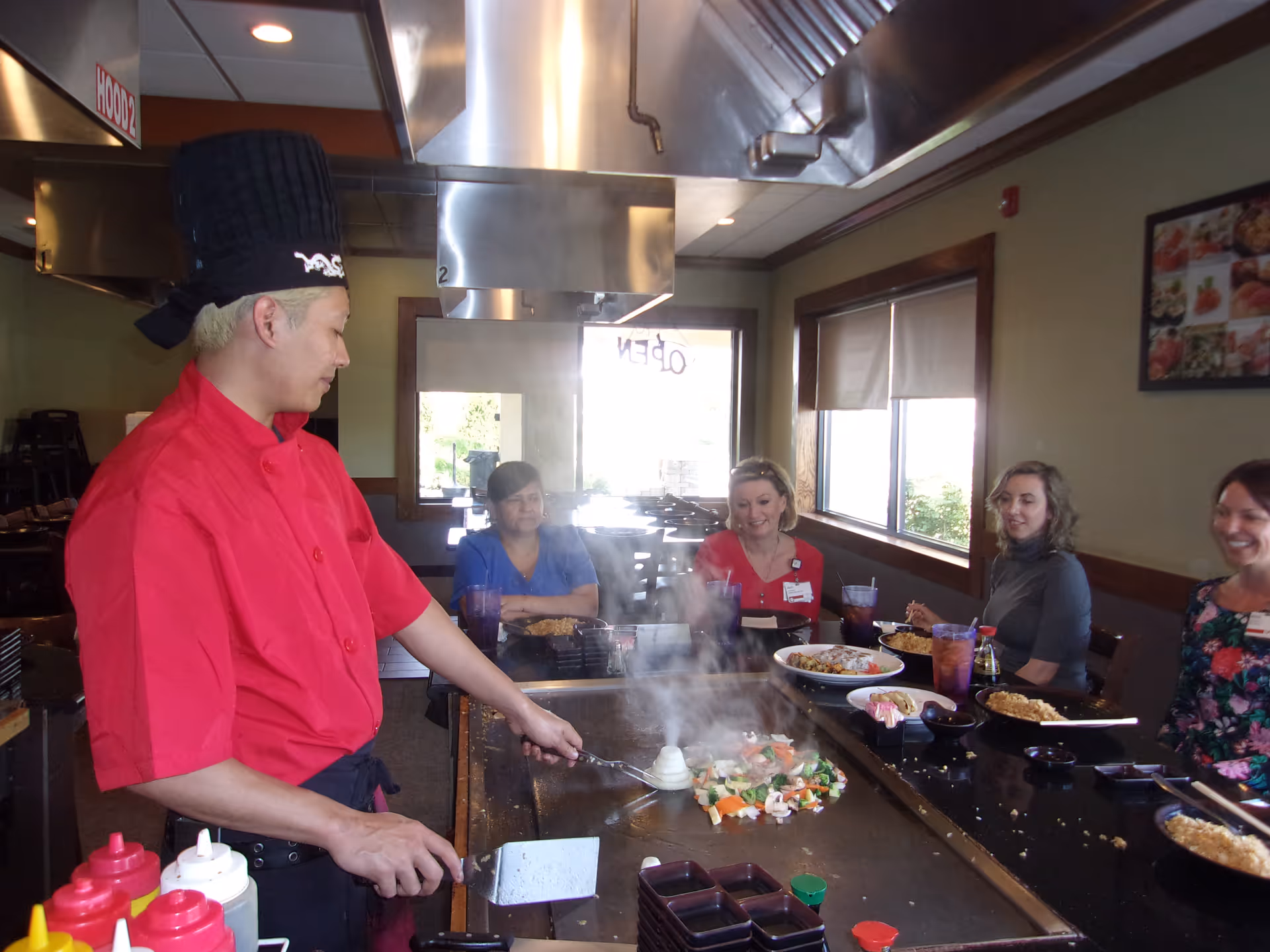 A chef wearing a red uniform and black chef hat is cooking vegetables and shrimp on a hibachi grill in a restaurant. Four women are seated around the grill, watching and smiling. The restaurant has large windows with blinds and framed pictures on the wall.
