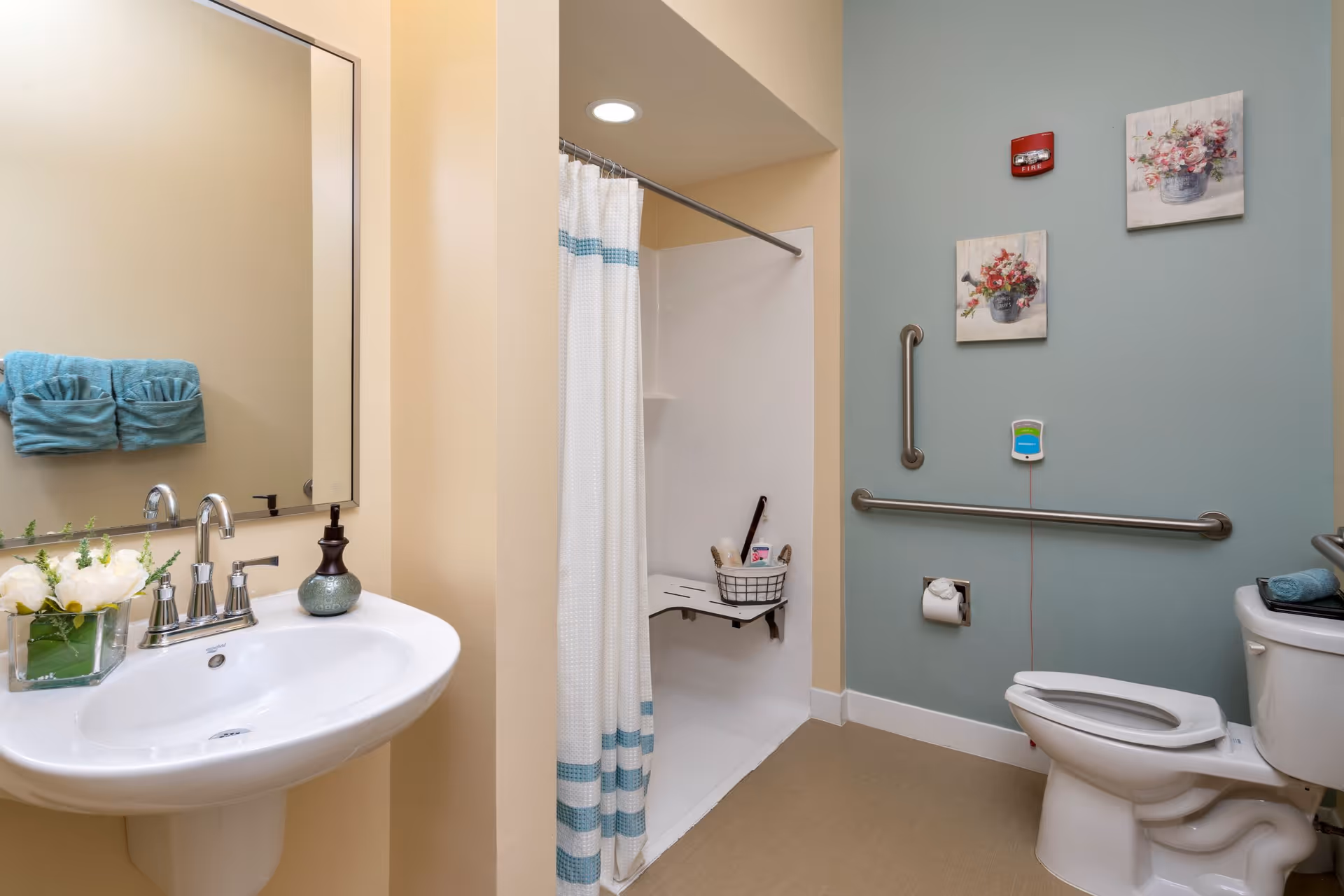 A clean and accessible bathroom featuring a white sink with a silver faucet, a large mirror above it, and a small vase with white flowers on the sink. There are two teal towels hanging on the wall. The bathroom includes a walk-in shower with a white curtain and a fold-down seat inside. The toilet has grab bars on the adjacent walls for support, and two floral paintings hang on the blue-green wall above the toilet paper holder and grab bars.