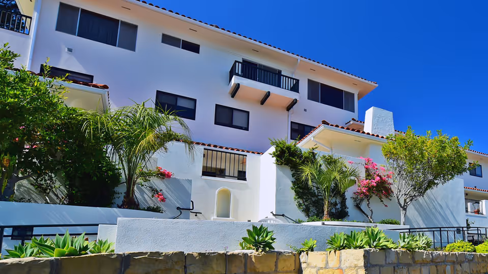 Exterior view of a white multi-story assisted living facility building with black-framed windows and small balconies. The building is surrounded by green plants, palm trees, and flowering bushes under a clear blue sky.