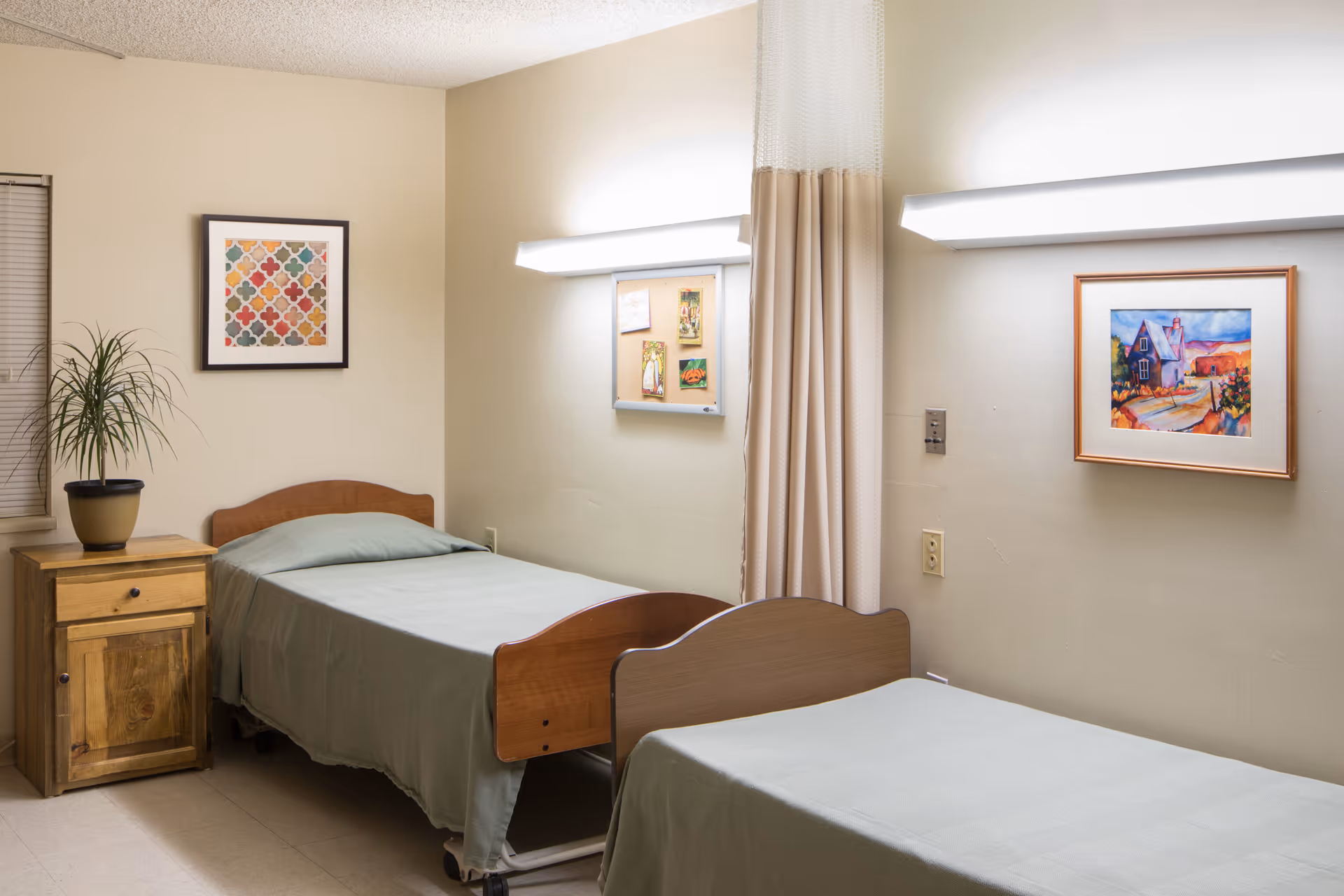 Two twin beds with green linens in a clinical patient room featuring a wooden nightstand, wall art, and a privacy curtain.