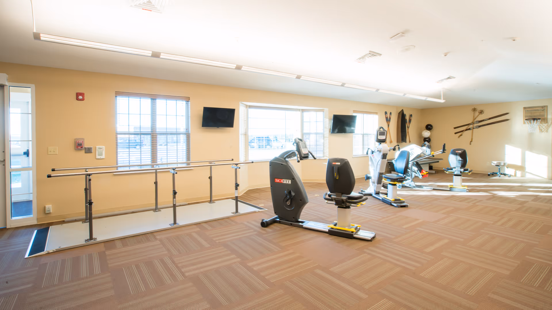A bright and spacious exercise room in a senior living facility with large windows letting in natural light. The room features exercise equipment including stationary bikes and a parallel bar setup for physical therapy. The walls are decorated with vintage skis and a basketball hoop.