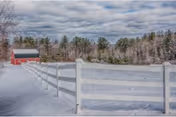 Snow-covered field with a white fence leading toward a red barn and a line of trees under a cloudy sky.